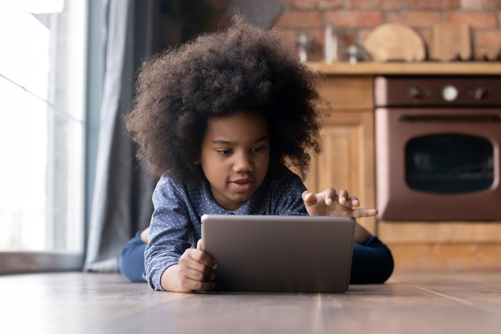 A young black girl with natural hair laying on the floor having screen time. She appears very engaged in her tablet.